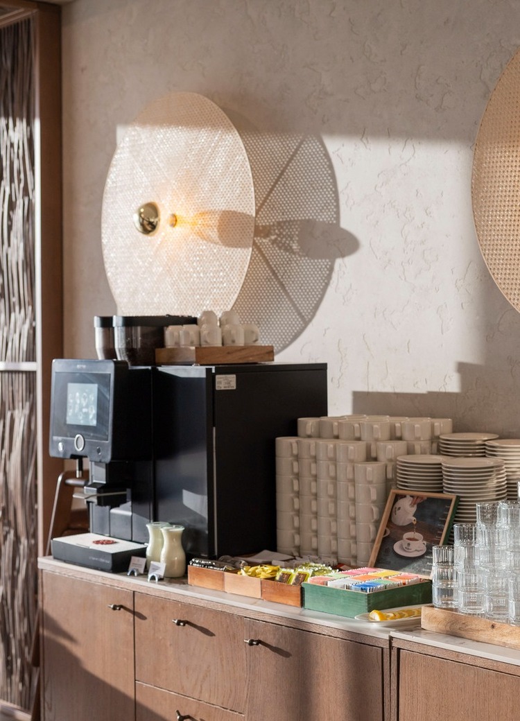 Selbstbedienungs-Kaffeezone im Hotel mit Kaffeeautomaten, gestapelten Tassen und Tellern, Gläsern, Teeauswahl auf Holztheke und warmem Wandlicht