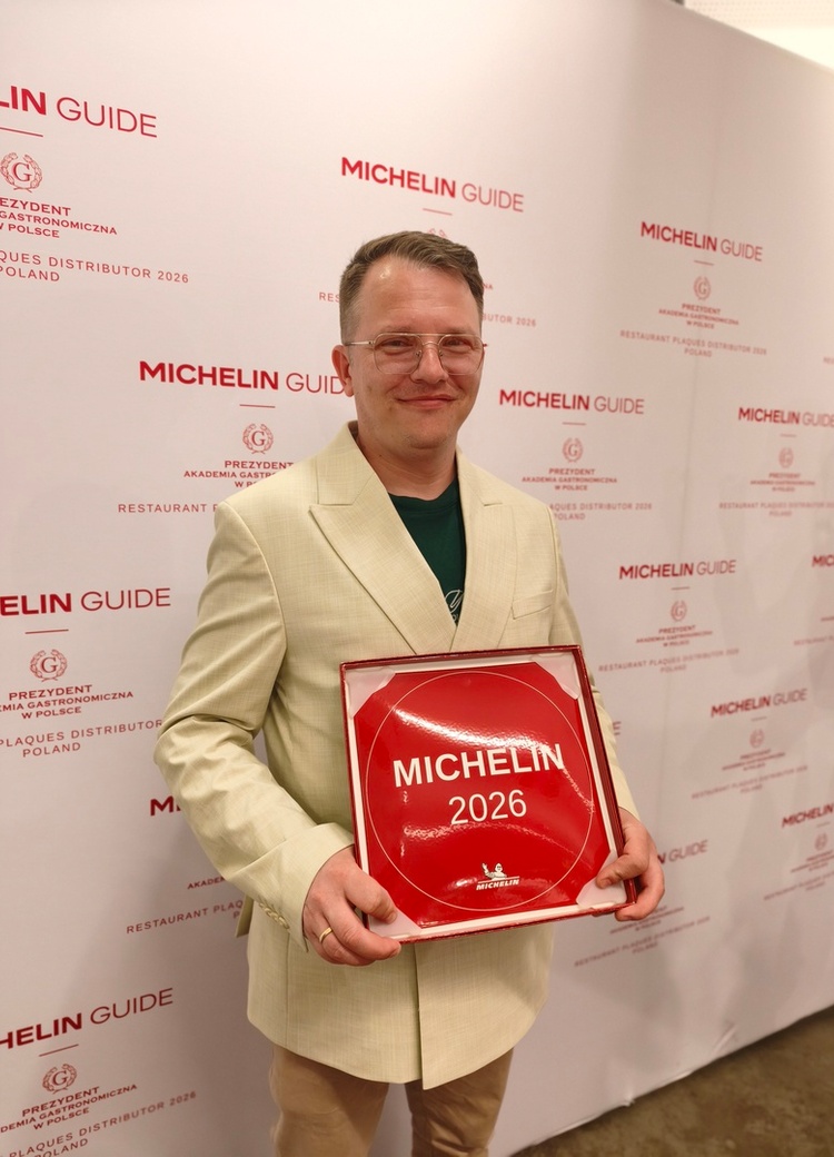 Osoba w jasnej marynarce trzyma czerwoną tablicę 'MICHELIN 2026' na tle baneru MICHELIN Guide