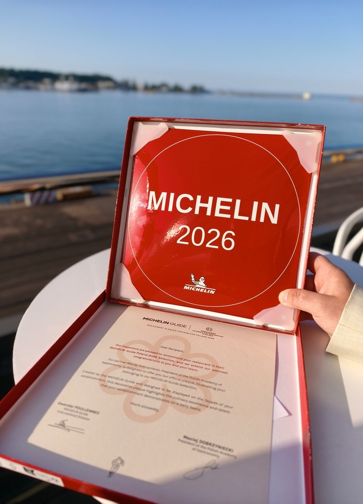 Czerwona tablica MICHELIN 2026 w otwartym pudełku z certyfikatem, dłoń trzymająca ją na nadmorskim tarasie.