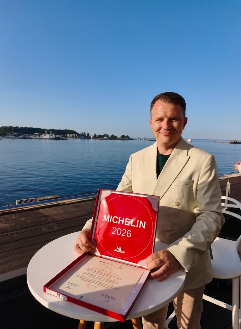 Mężczyzna w beżowym garniturze trzyma nagrodę Michelin 2026 na tarasie przy wodzie z widokiem na spokojne morze