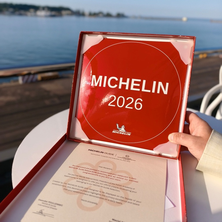 Czerwona tablica MICHELIN 2026 w otwartym pudełku z certyfikatem, dłoń trzymająca ją na nadmorskim tarasie.