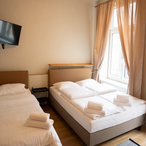 Pokój hotelowy z dwiema pojedynczymi łóżkami, jasnymi zasłonami i telewizorem na ścianie, złożone ręczniki na łóżkach.