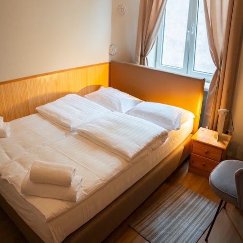 Przytulny pokój z podwójnym łóżkiem, białą pościelą, drewnianą szafką nocną i szarym fotelem przy oknie z zasłonami.