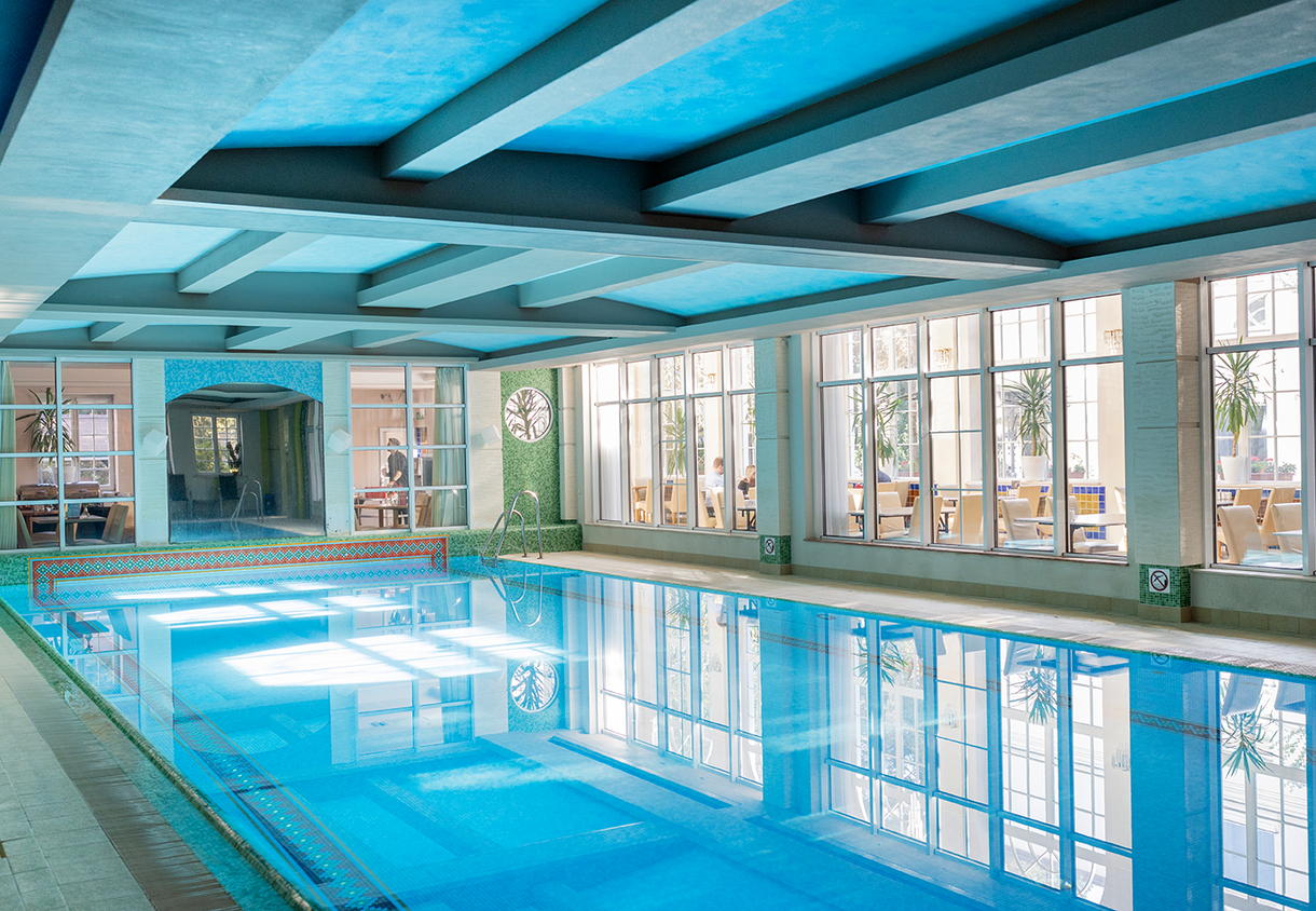 Ogrzewany kryty basen hotelowy z niebieskim sufitem kasetonowym, dużymi oknami i widoczną jadalnią za szybą.