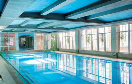 Jasny kryty basen hotelowy z niebieskim stropem i belkami, dużymi oknami, roślinami w doniczkach, mozaikową krawędzią i stalową drabinką.