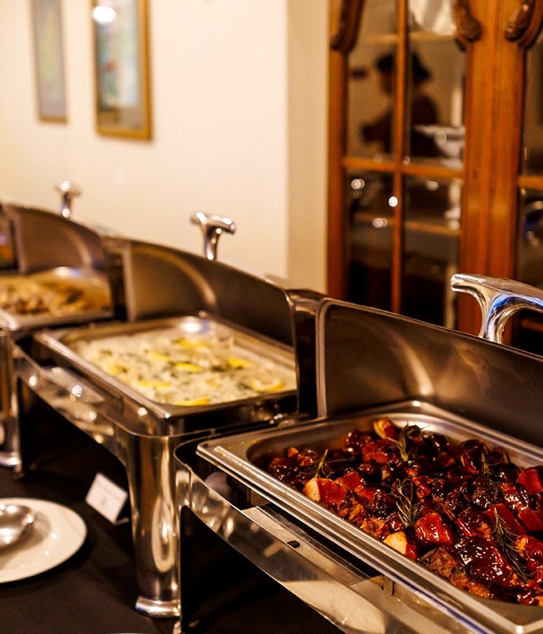Bufet hotelowy z podgrzewaczami ze stali, serwujący glazurowane pieczenie w sosie, kremową zapiekankę i gorące dodatki, talerze i łyżki.