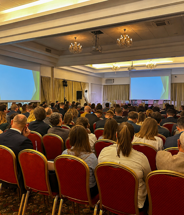 Sala konferencyjna hotelu przygotowana na wydarzenie biznesowe, rzędy czerwonych krzeseł, dwa ekrany projekcyjne, scena i żyrandole.