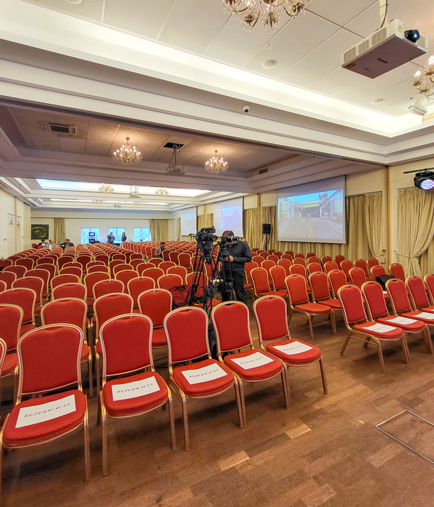 Elegancka sala konferencyjna hotelu z rzędami czerwonych krzeseł, żyrandolami, ekranami projekcyjnymi, kamerą i zarezerwowanymi miejscami.