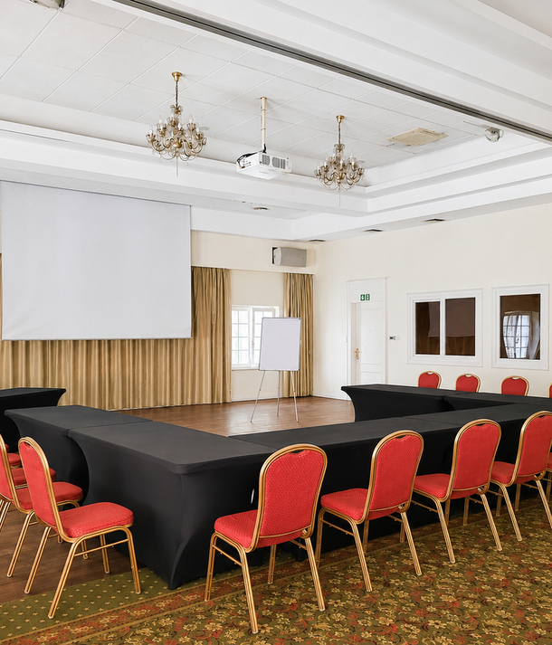 Sala konferencyjna w układzie podkowy z czarnymi obrusami, czerwonymi krzesłami, projektorem i ekranem, flipchartem, żyrandolami i beżowymi zasłonami.