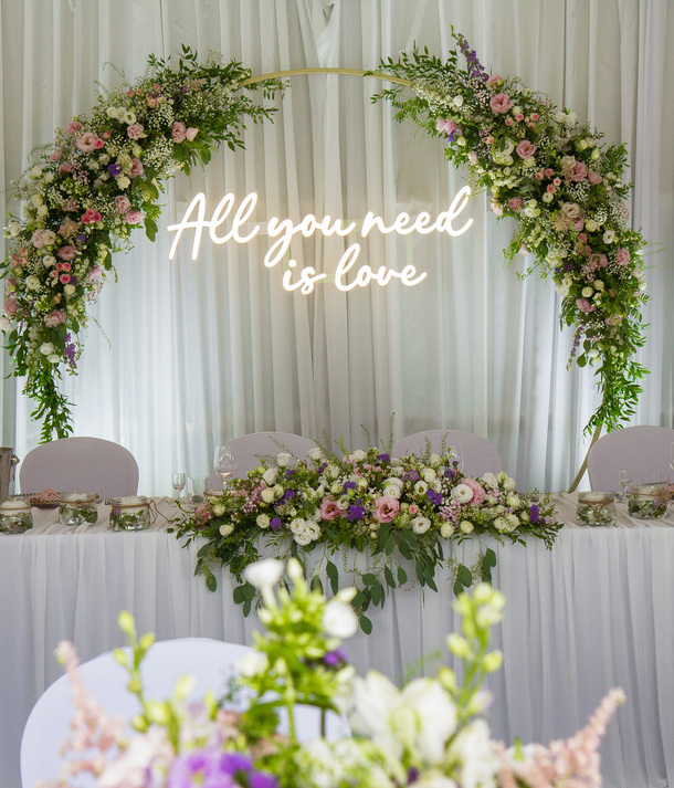 Stół główny wesela z pastelowym łukiem kwiatowym, neonowym napisem 'All you need is love', białą zasłoną i eleganckimi kompozycjami.