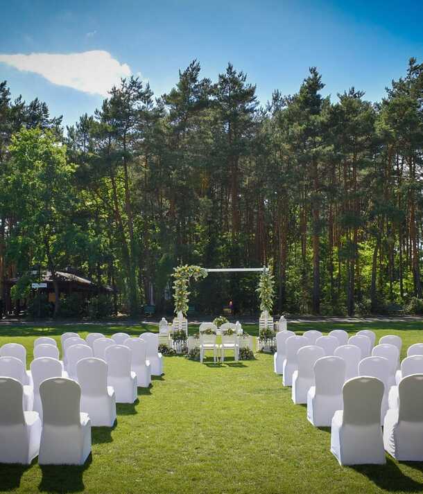 Plenerowa ceremonia ślubna na zadbanym trawniku: rzędy krzeseł w białych pokrowcach tworzą aleję do kwiatowej bramy, w tle sosnowy bór.