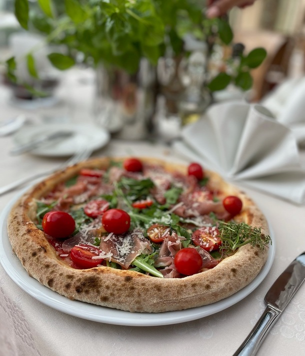 Pizza z prosciutto, rukolą, pomidorkami koktajlowymi i tartym parmezanem na talerzu, podana na lnianym obrusie.
