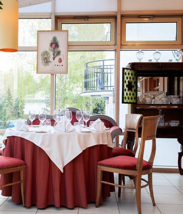 Przyszklony, słoneczny kącik restauracyjny z okrągłym stołem w bordowej spódnicy i białym obrusem, złożonymi serwetkami, kieliszkami i witryną