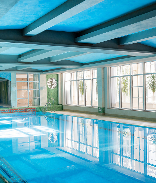 Jasny kryty basen hotelowy z niebieskim stropem i belkami, dużymi oknami, roślinami w doniczkach, mozaikową krawędzią i stalową drabinką.