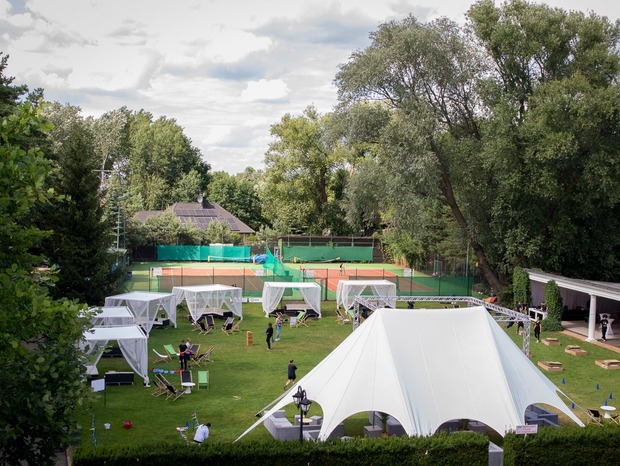 Trawnik hotelowy przygotowany na event z dużym białym pawilonem, odsłoniętymi kabinami, leżakami i kortami tenisowymi w tle.