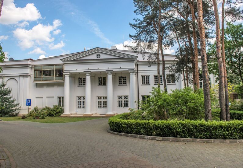 Neoklasycystyczna biała elewacja hotelu z wysokimi kolumnami, eleganckim wejściem, sosnami i zadbanym podjazdem