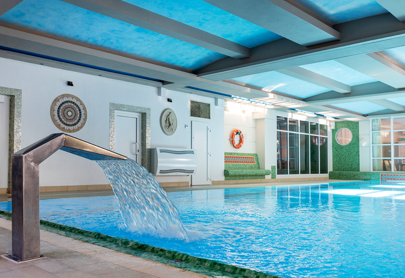 Kryty basen hotelowy z metalową kaskadą wodną, mozaikami, siedziskami i kołem ratunkowym, subtelne niebieskie oświetlenie
