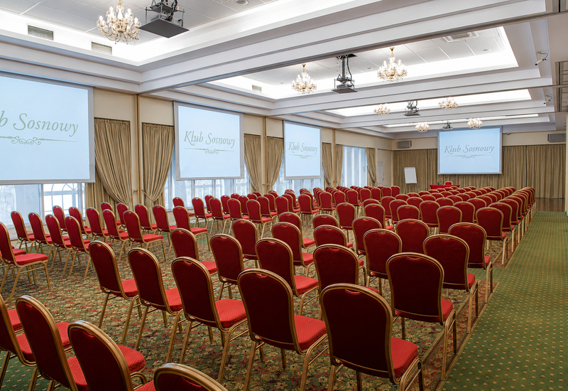 Sala konferencyjna z czerwonymi krzesłami, ekranami z napisem 'Klub Sosnowy', żyrandolami, wzorzystym dywanem i zasłonami
