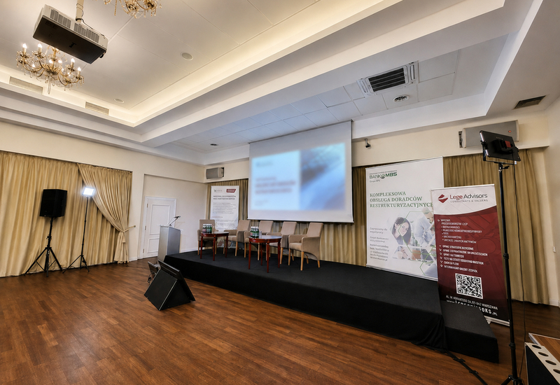 Sala konferencyjna hotelu z podwyższeniem, ekranem projekcyjnym, czterema fotelami panelowymi, mównicą i żyrandolami