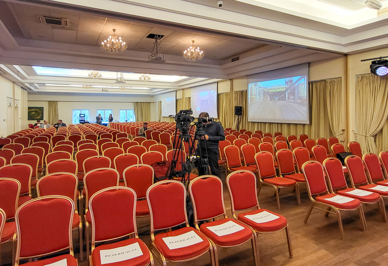 Elegancka sala konferencyjna hotelu z rzędami czerwonych krzeseł, żyrandolami, ekranami projekcyjnymi, kamerą i zarezerwowanymi miejscami.