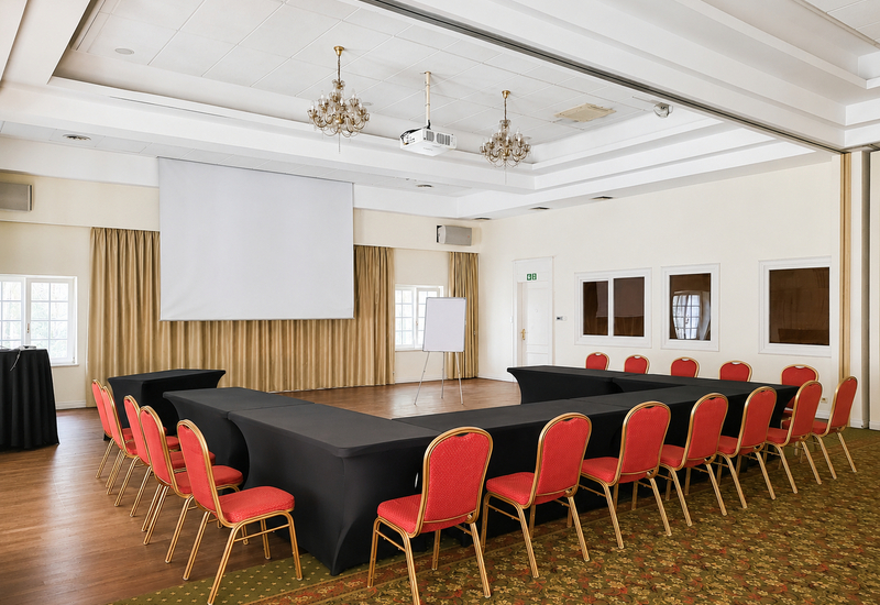 Sala konferencyjna w układzie podkowy z czarnymi obrusami, czerwonymi krzesłami, projektorem i ekranem, flipchartem, żyrandolami i beżowymi zasłonami.