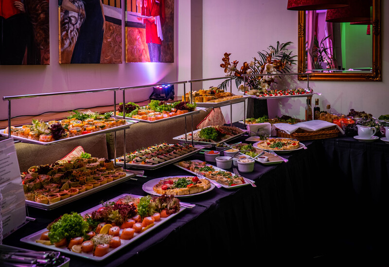 Elegancki bufet hotelowy z kanapkami, wędzonym łososiem, wędlinami, sałatkami i tartami na piętrowych półmiskach w ciepłym świetle.