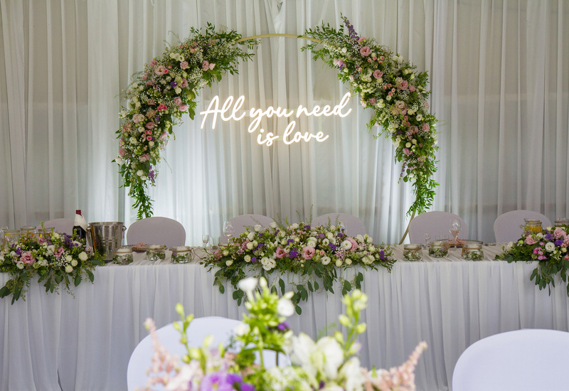 Stół główny wesela z pastelowym łukiem kwiatowym, neonowym napisem 'All you need is love', białą zasłoną i eleganckimi kompozycjami.