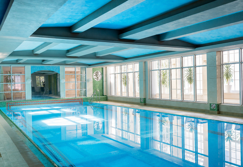 Jasny kryty basen hotelowy z niebieskim stropem i belkami, dużymi oknami, roślinami w doniczkach, mozaikową krawędzią i stalową drabinką.