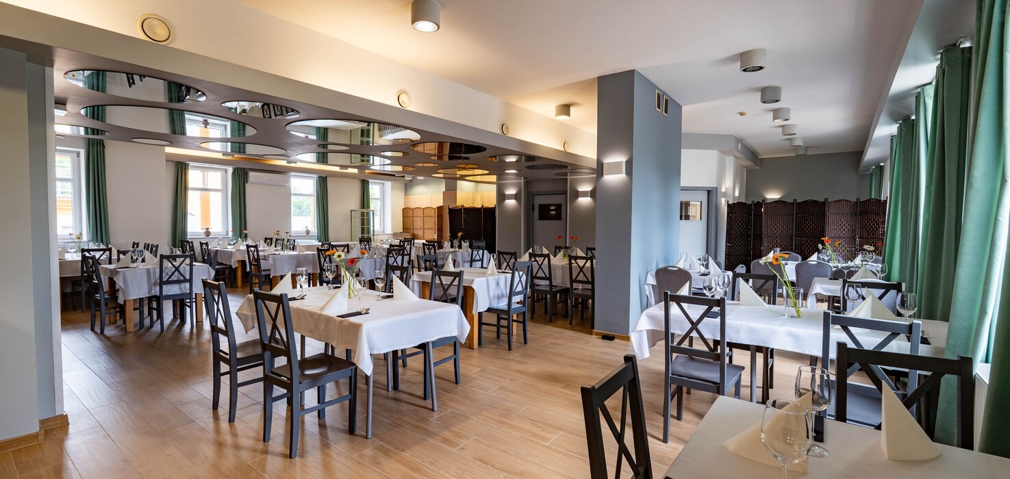 Spacious hotel restaurant with white tablecloths, place settings and wine glasses, wooden chairs, green curtains and round ceiling panels