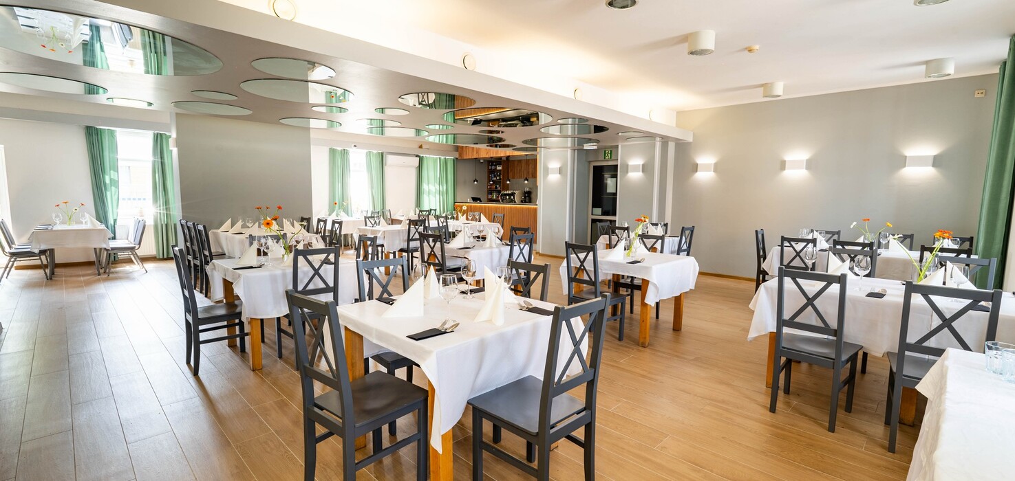 Bright hotel restaurant with white tablecloths, wooden floor, green curtains, mirrored round panels and bar.