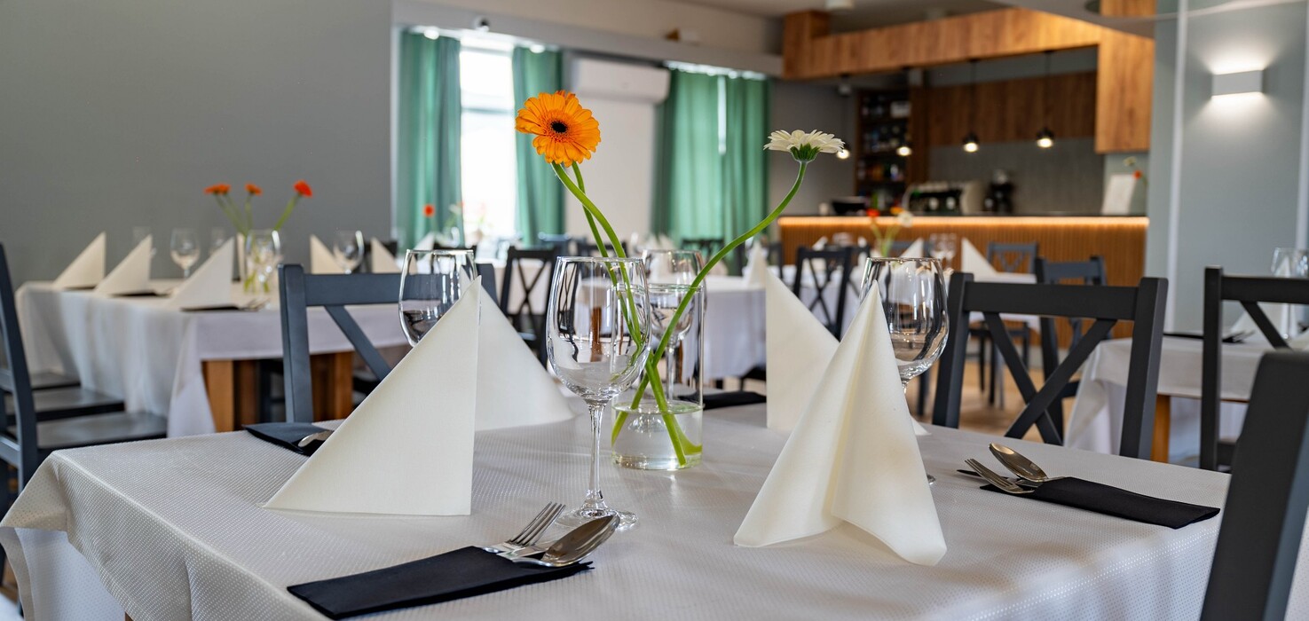 Hotel restaurant with white tablecloths, folded napkins, wine glasses and gerberas in vases; modern wood bar and green curtains.