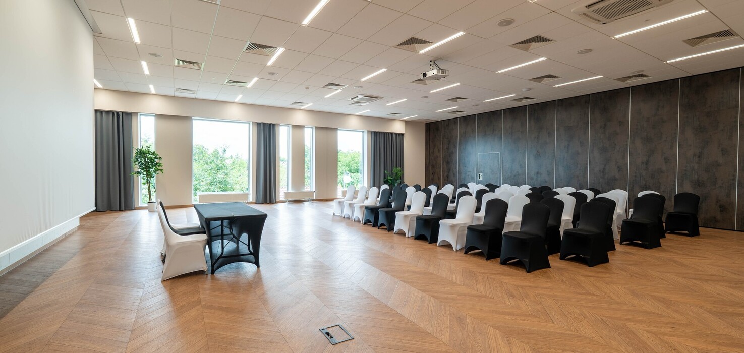 Jasna sala konferencyjna w hotelu z rzędami czarno-białych pokrowców, projektorem sufitowym, ekranem, drewnianą podłogą i dużymi oknami.