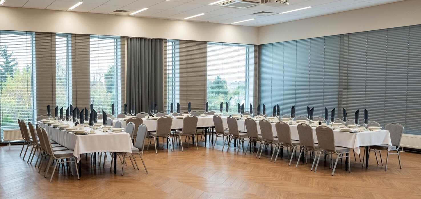 Sala bankietowa z długimi stołami przykrytymi białymi obrusami, czarne złożone serwetki, szkło, projektor sufitowy, duże okna