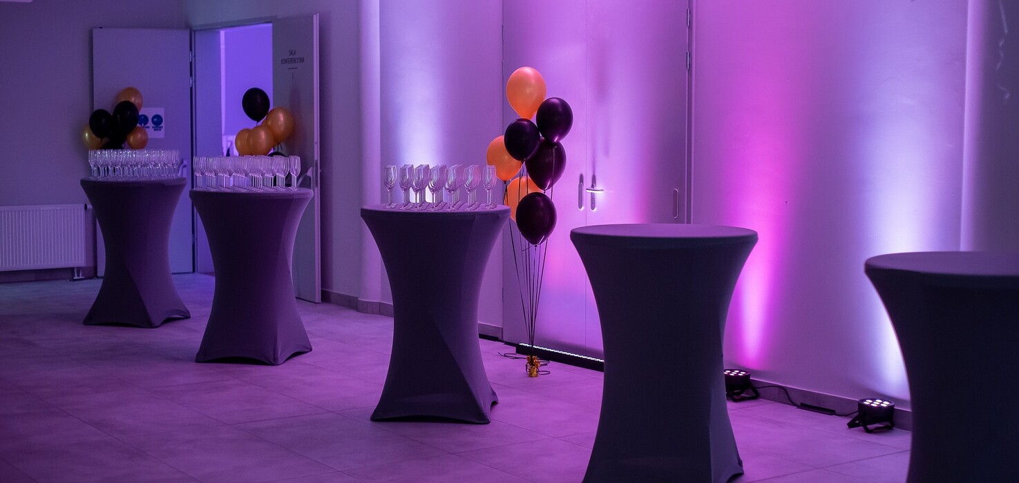 Sala eventowa na koktajl: wysokie stoły w pokrowcach, kieliszki i czarno-pomarańczowe balony w fioletowym świetle.
