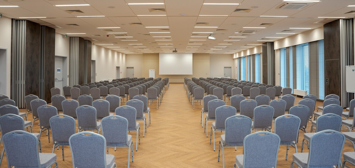Przestronna nowoczesna sala konferencyjna z rzędami szarych krzeseł, ekranem, drewnianą podłogą i oknami z żaluzjami