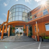 Hotelowe wejście z przeszklonym, zaokrąglonym atrium, pomarańczową elewacją, kolumnami, schodami i donicami.