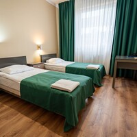 Pokój z dwoma łóżkami pojedynczymi, zielonymi narzutami, złożonymi ręcznikami, stolikami nocnymi, biurkiem i zasłoniętym oknem.