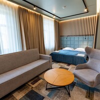 Nowoczesny pokój hotelowy z łóżkiem king‑size w niebieskiej narzucie, tapicerowany zagłówek z listwami, sofą, fotelem, okrągłym stolikiem i firanami.