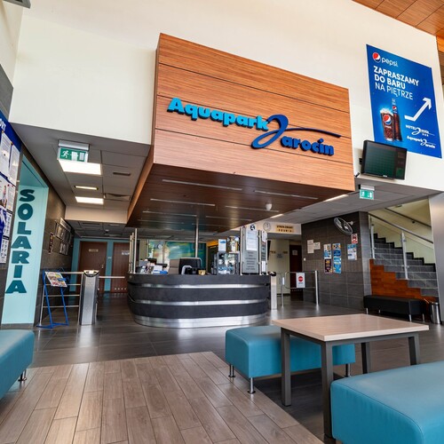 Aquapark reception with a rounded black desk, wooden 'Aquapark Jarocin' sign, turquoise seats, stairs and information board.