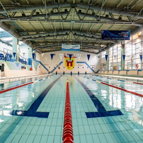 Indoor training pool with lanes and starting blocks, grandstands, large windows and exposed ceiling pipes.