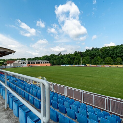 Open stands, one covered section, blue seats, well-kept pitch, changing-room building, woodland backdrop