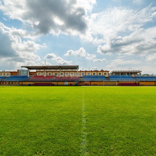 Green football pitch before a covered stand with red and blue seats under a cloudy sky