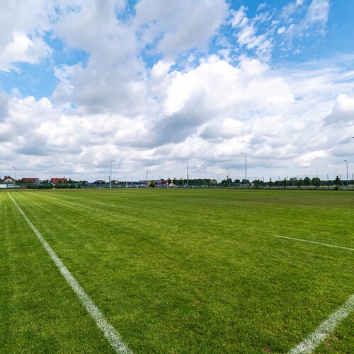 Well-maintained grass pitch with white lines, lighting and a small pavilion by the fence under a cloudy sky