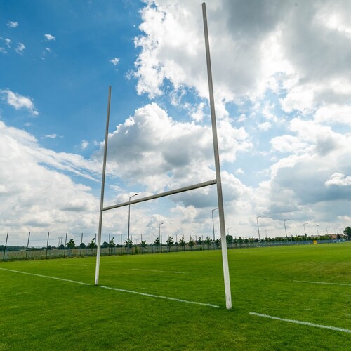 Rugby goalposts on a well-kept green pitch under a hazy blue sky, with fencing and lights.