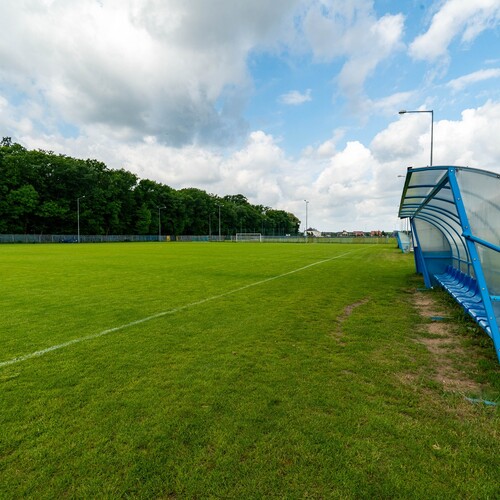 Football pitch with a well-kept lawn, blue covered team benches, distant goal and trees under clouds.
