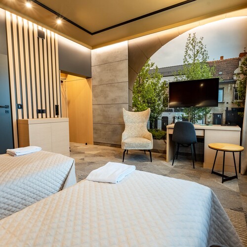 Modern twin room with quilted beds and towels, wood wall, comfortable armchair, desk with TV and a window view.