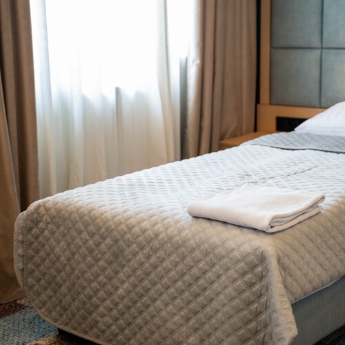Bright hotel room with quilted bedspread, folded towels on the bed, upholstered headboard and sheer curtains