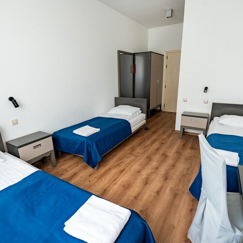 Bright minimalist triple room with three single beds, blue bedspreads, bedside tables, lamps and wardrobe
