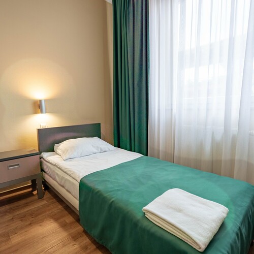 Room with two single beds, green bedspreads, bedside table with wall lamp, sheer curtain and folded towels