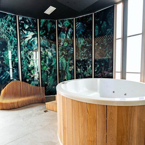 Private spa with round jacuzzi in wooden casing, wavy wooden lounger and illuminated glass wall with botanical motif.